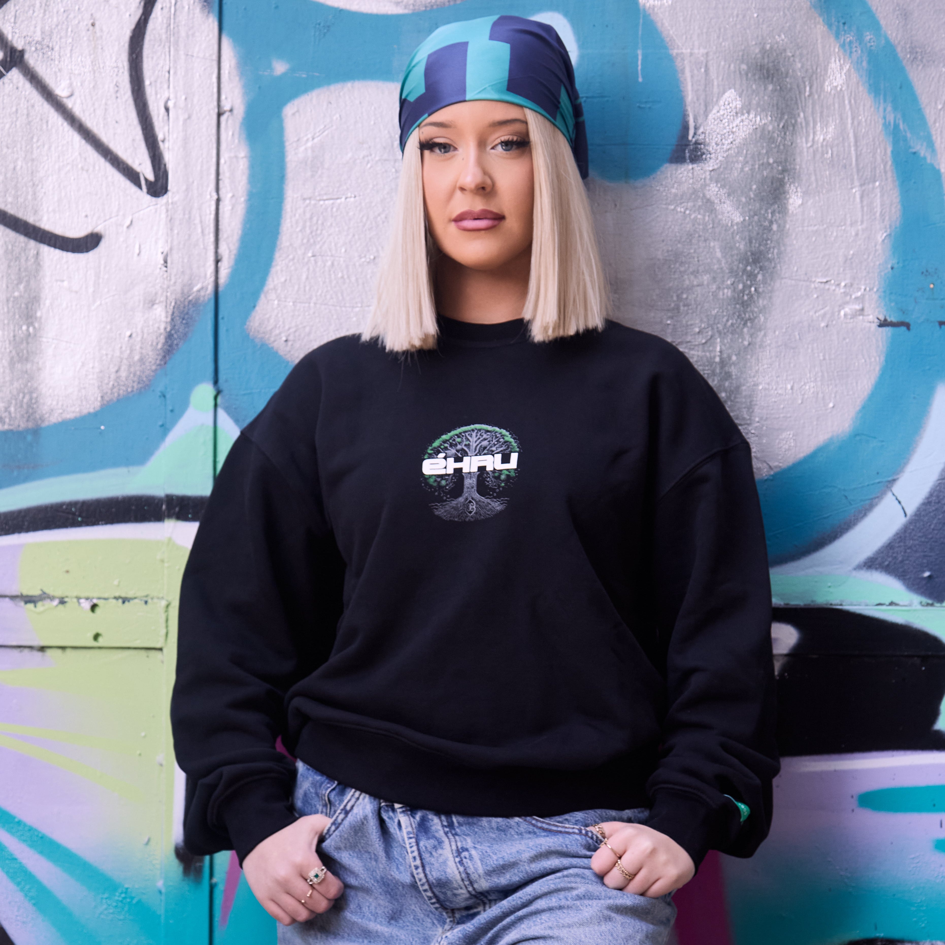 Ella is wearing an Ancestral Memory sweatshirt with a logo and light blue jeans standing against a graffiti-covered wall.